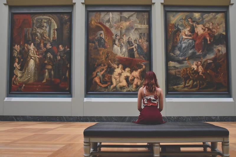 Hier sehen Sie eine(n)Besucher:in in einem Museum vor einem dreiteiligen Bild (Triptychon) sitzen