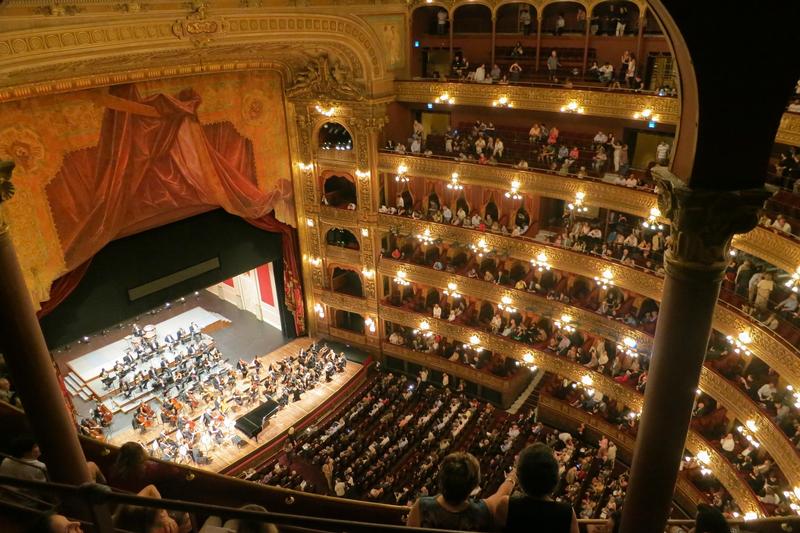 Hier sehen Sie einen Theaterraum mit Zuschauern auf ihren Sitzen und einem Orchester auf der Bühne